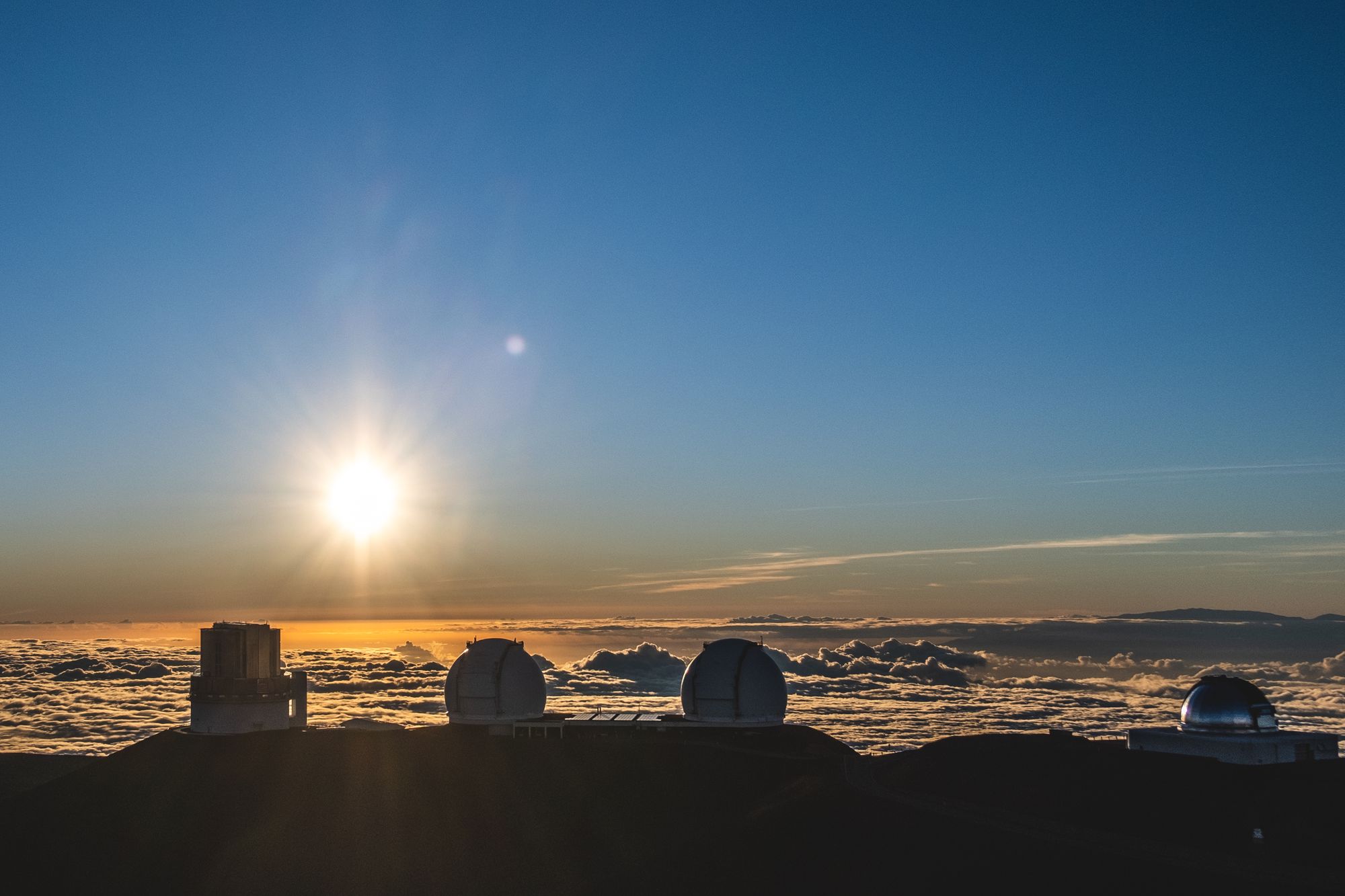 A photo of the sun setting on the horizon, with clouds directly in front. In the foreground are 4 large telescope facilities used for night sky gazing and other astrophysics research.